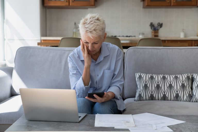 Worried middle aged woman at home reviewing paper bills and a laptop, hand to her head