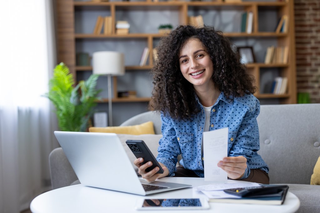 Smiling woman at home holding a paper bill and using a laptop and phone to manage her budget