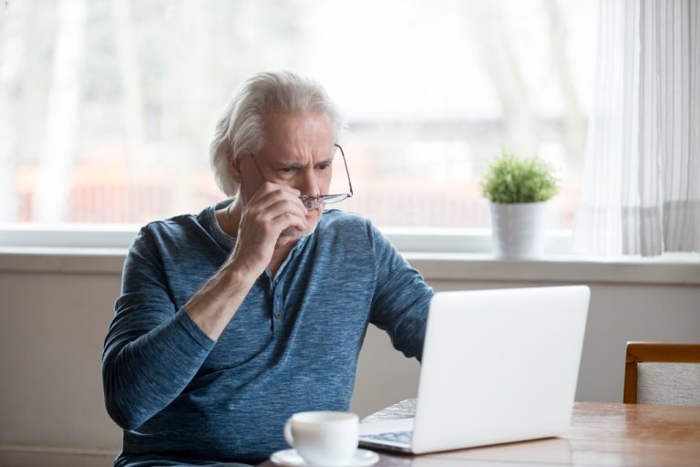 Worried older man at home taking off his glasses to look closely at his laptop, reacting to unexpected financial news