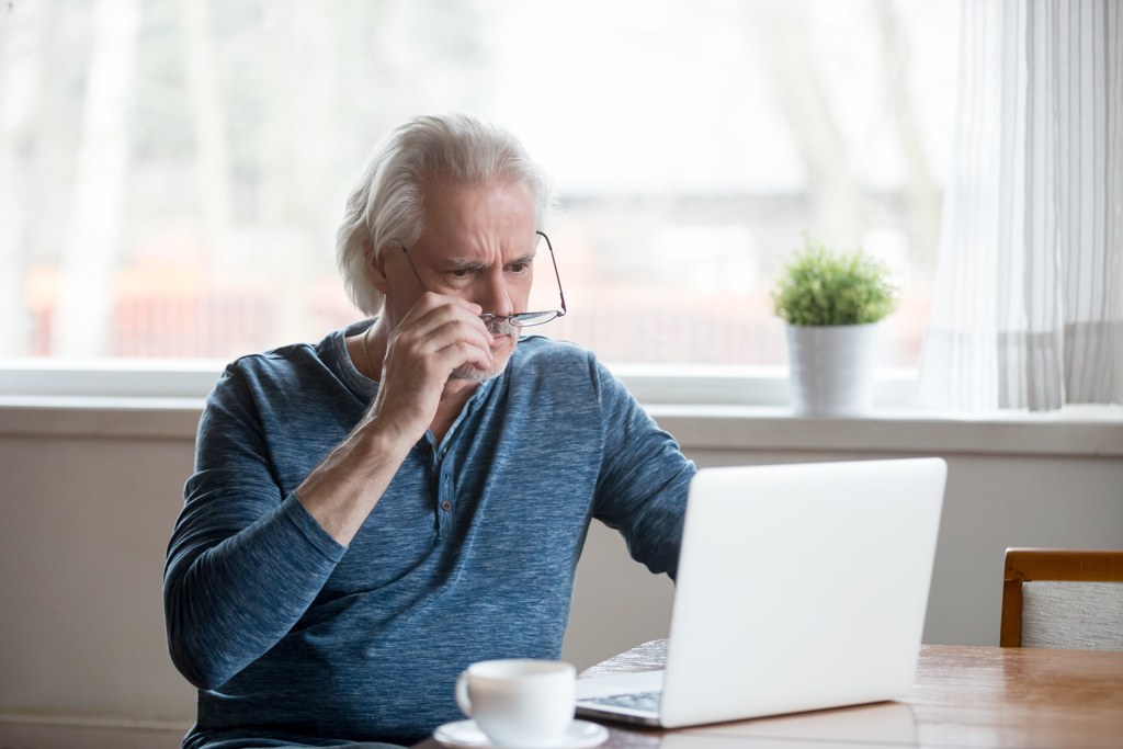 Worried older man at home taking off his glasses to look closely at his laptop, reacting to unexpected financial news